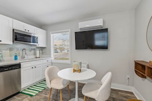 Una cocina moderna con armarios blancos, lavavajillas, microondas y horno empotrado. Hay una mesa redonda blanca con dos sillas y un pequeño adorno encima. Un televisor de pantalla plana está montado en la pared sobre la mesa.