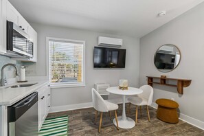 Cocina compacta con lavavajillas, una ventana con persianas, televisor montado en la pared, espejo redondo y pequeño comedor con mesa y sillas blancas.