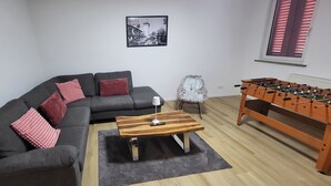 Olohuoneessa on sohva, sohvapöytä, lamppu, pöytäjalkapallopeli ja seinällä oleva kehystetty kuva.