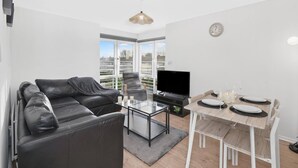 Una sala de estar moderna con un sofá de cuero negro, una mesa de centro de vidrio, un televisor y una mesa de comedor preparada para cuatro personas.