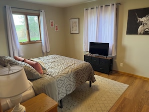 A bedroom with a bed, a TV on a stand, a lamp, and a window with curtains.
