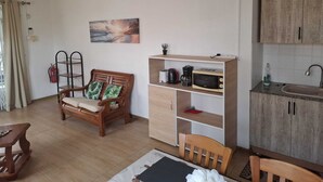 Een compacte leefruimte met een kitchenette, een eethoek met een houten tafel, een bank met kussens, een plafondventilator en een schilderij aan de muur.