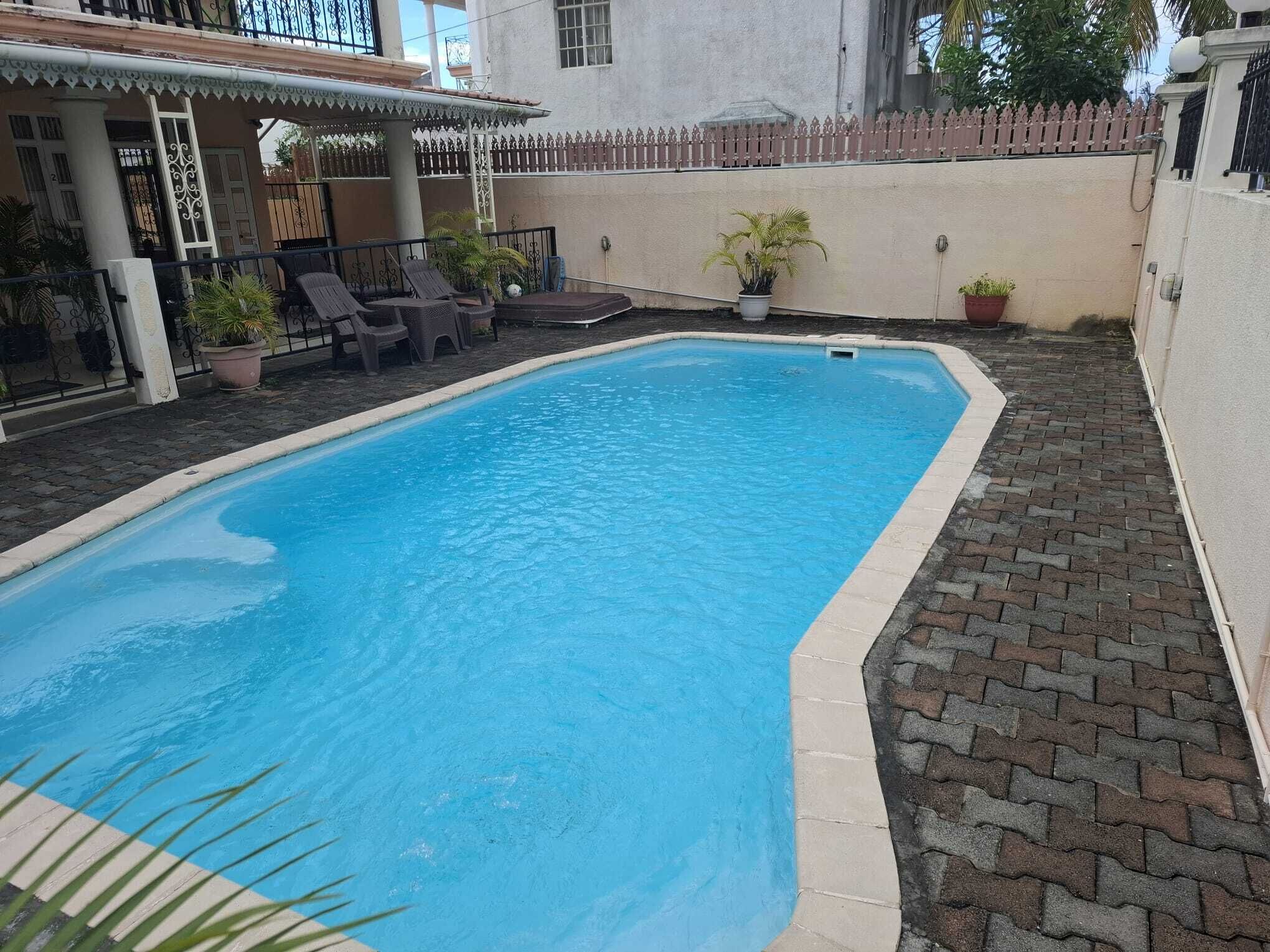 A rectangular swimming pool with a tiled edge, surrounded by a brick patio and a white wall.
