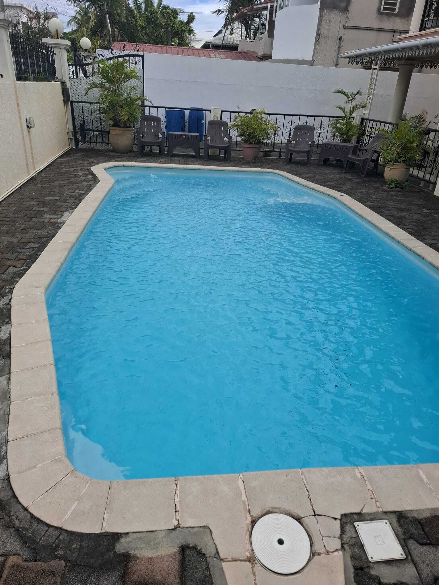 Una piscina rectangular con borde de azulejos, rodeada por una zona pavimentada con sillas y plantas en macetas.