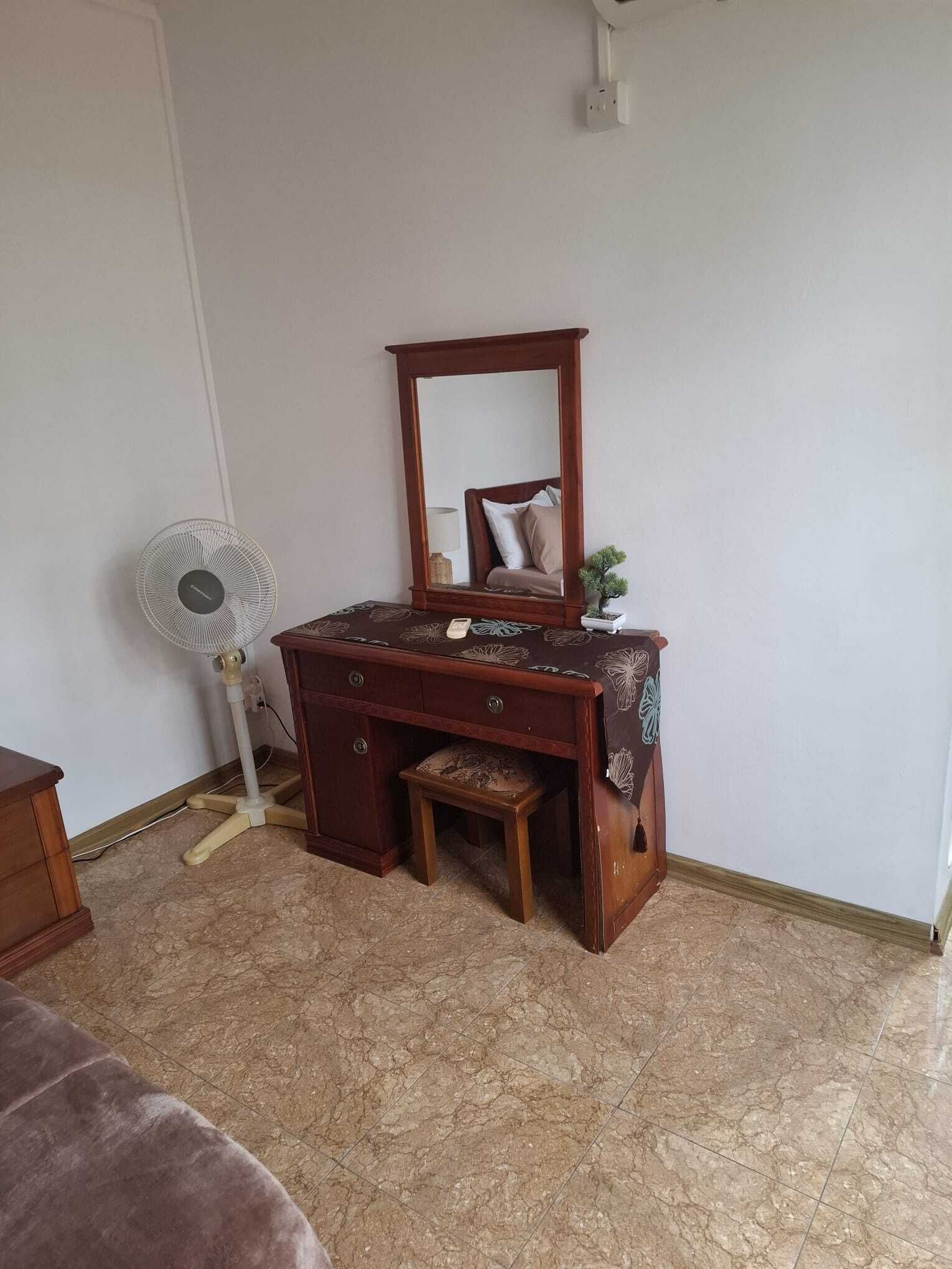 A room with a wooden dresser, a mirror, a small stool, a fan, and a potted plant.