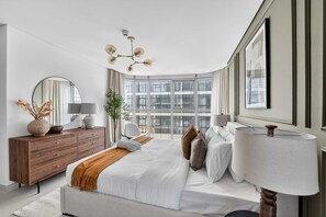 A modern bedroom with a large bed, a dresser, a mirror, a chandelier, and large windows with a city view. - Silkhaus Vacation Homes Elegant 2BR With Maid Room in Radiant Heights (Abu Dhabi)