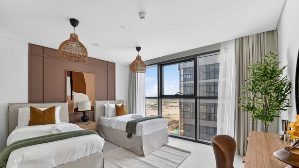 Une chambre d’hôtel moderne équipée de deux lits, d’un bureau, d’une chaise, d’une grande fenêtre donnant sur la ville et de deux luminaires suspendus.