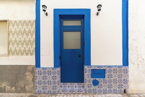 Eine blaue Tür mit einer Milchglasscheibe, ein kleines Fenster darüber und eine Wand mit gemusterten Fliesen.