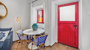 Een kamer met een ronde tafel, twee blauw gestreepte stoelen, een blauwgroene vaas, een rode deur en een witte muur met een spiegel.