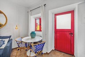 Ein Zimmer mit einem runden Tisch, zwei stuhlförmigen Sesseln mit blauem Muster, einer türkisfarbenen Vase, einer roten Tür und einer weißen Wand mit einem Spiegel.