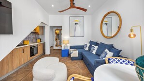 Un soggiorno moderno con una cucina, un divano blu, una poltrona bianca, una ventola a soffitto in legno e un grande specchio.