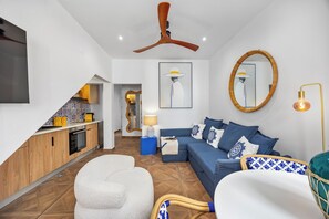 Um espaço de estar moderno com cozinha, um sofá azul, uma poltrona branca, um ventilador de teto de madeira e um espelho grande.