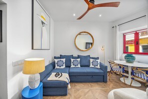 Een moderne woonkamer met een bank, een ronde spiegel, een plafondventilator en een eethoek met een tafel en stoelen.