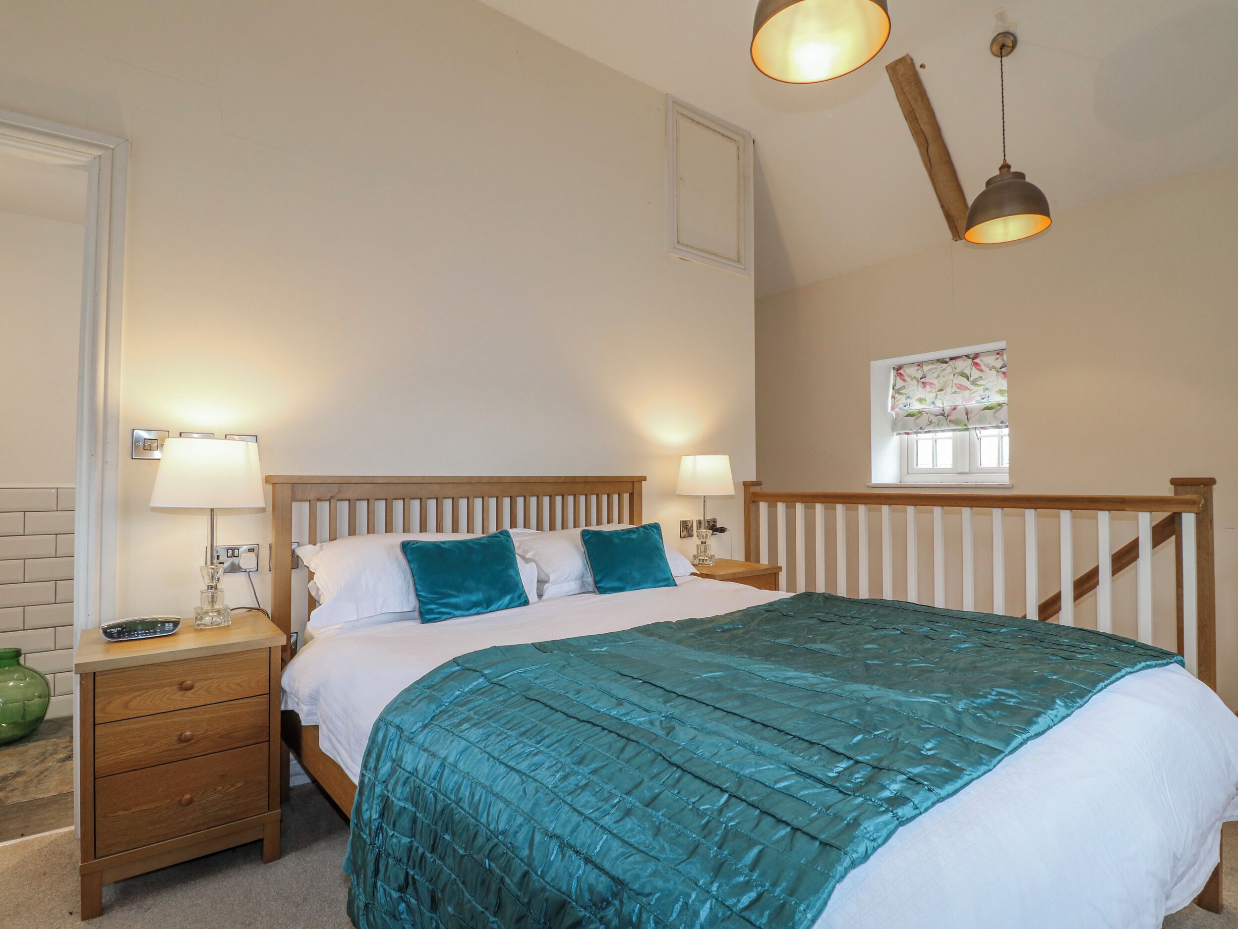 A bedroom with a bed, two bedside tables, a nightstand, a window with curtains, and a wooden ceiling beam.