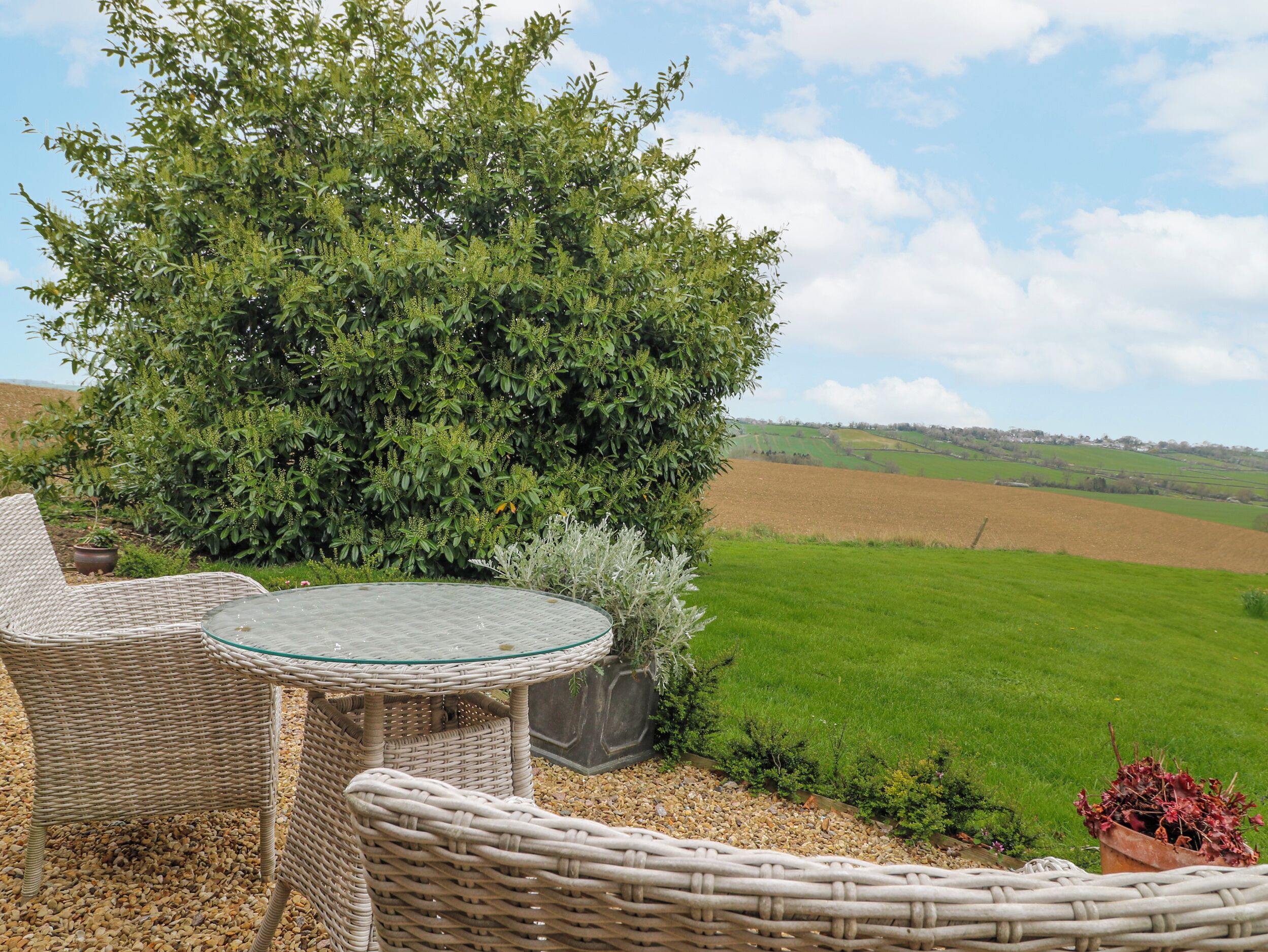 A wicker outdoor seating area with a glass table, surrounded by greenery and a view of a green field.