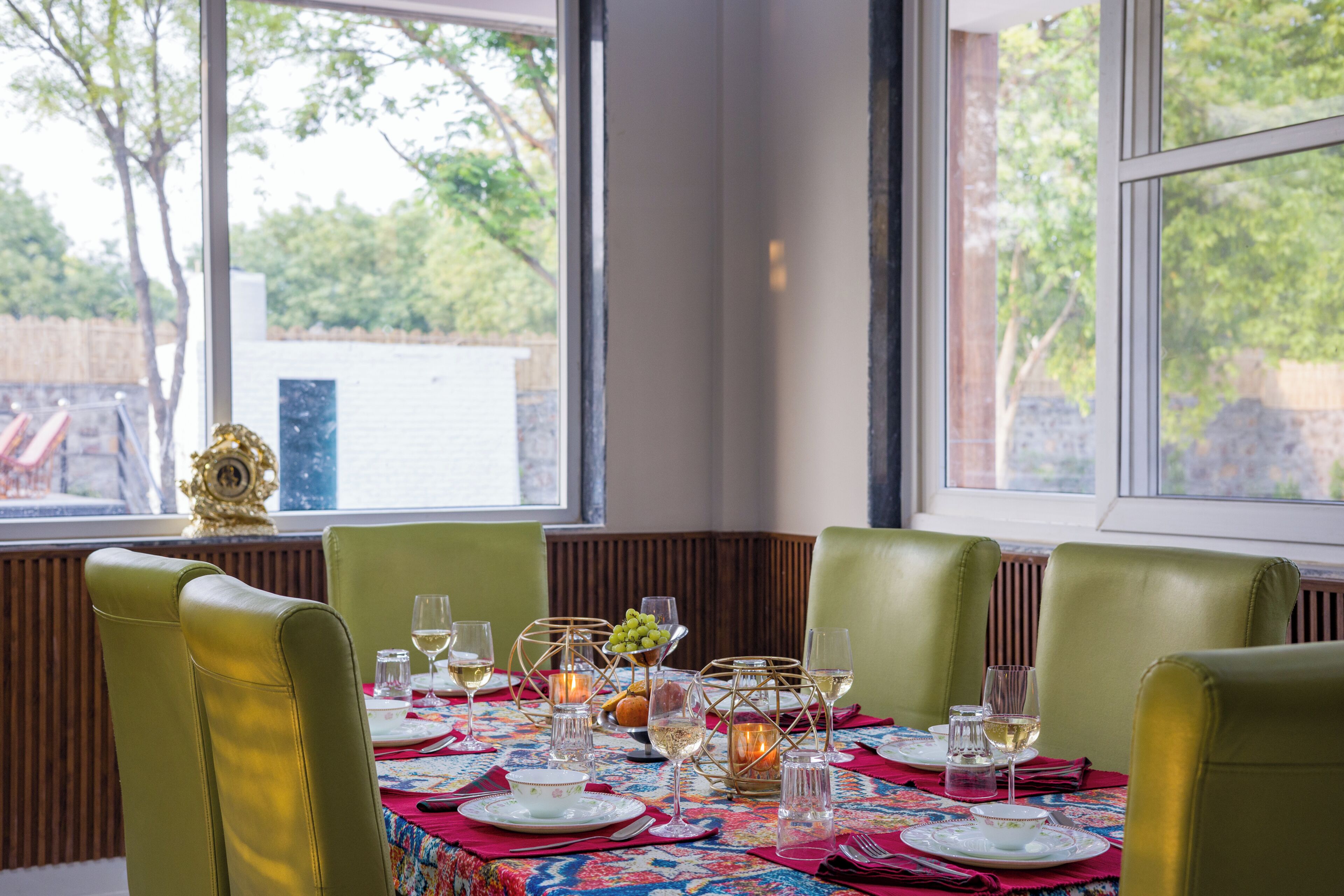 A dining table set for six with green chairs, a patterned tablecloth, and a view of trees and a building outside.
