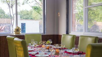 A dining table set for six with green chairs, a patterned tablecloth, and a view of trees and a building outside.