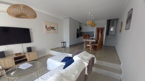 Ein Wohnzimmer mit einer Couch, einem Fernseher und Lautsprechern, eine Essecke mit Stühlen und einem Tisch sowie eine Küche im Hintergrund.