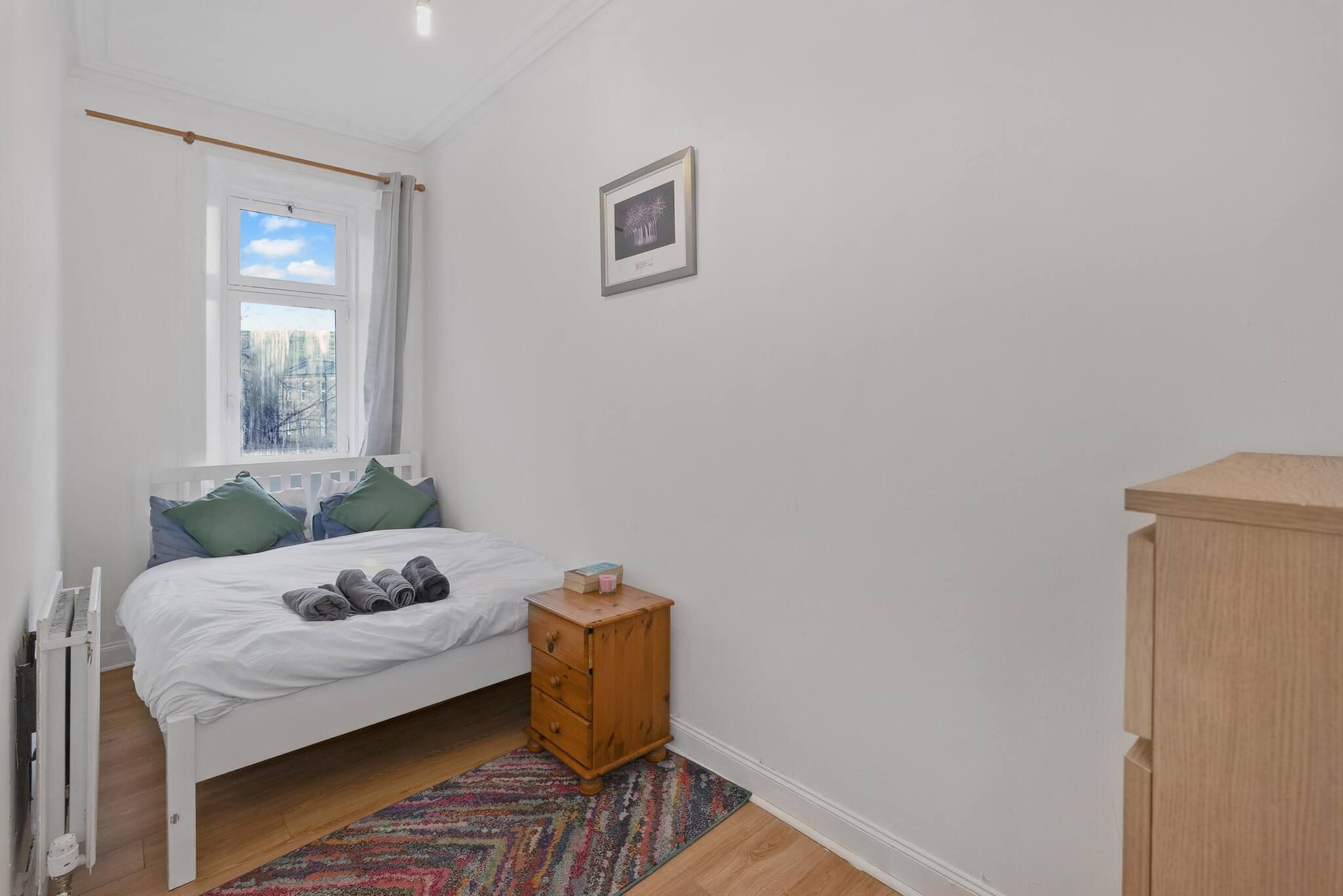 A bedroom with a bed, a wooden nightstand, a radiator, a window with curtains, and a picture on the wall.