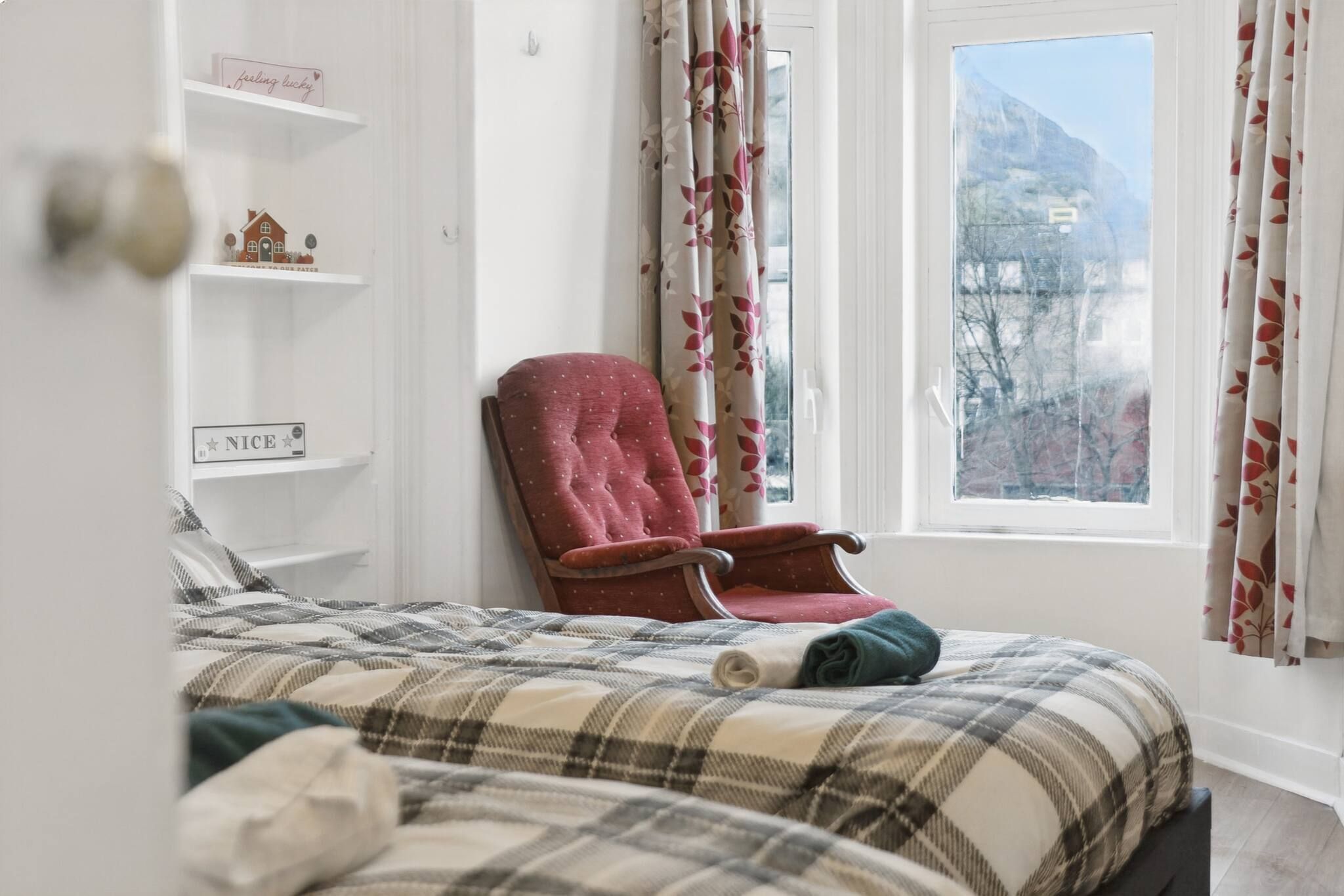 Une chambre à coucher avec un lit, un fauteuil rouge, une fenêtre donnant sur une montagne et des étagères ornées d’objets décoratifs.