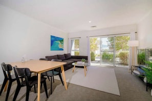 A modern living room with a sofa, a dining table with chairs, a coffee table, and a large window with a view of greenery. - Bright 2BDR Apt with Pool Access Williamstown (Williamstown)
