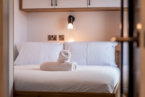 A neatly made bed with a rolled towel, white bedding, and a wall-mounted light fixture. - Marine Place | Flat 14 (St Andrews)