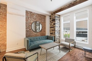 A room with a brick wall, a round mirror, a blue sofa, a patterned armchair, a wooden coffee table, and a window with a view. - Meramont Hotel Centre Ville (Montréal)