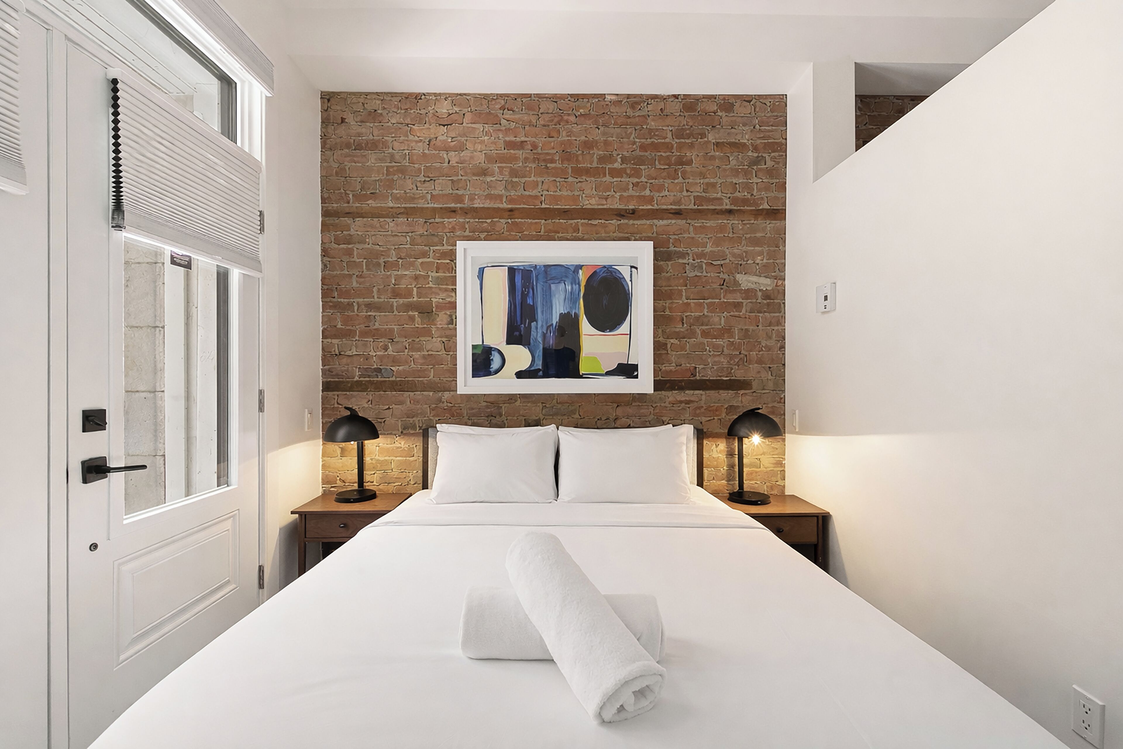 Une chambre d’hôtel avec un lit, un mur en briques, un tableau abstrait encadré et deux lampes de chevet.