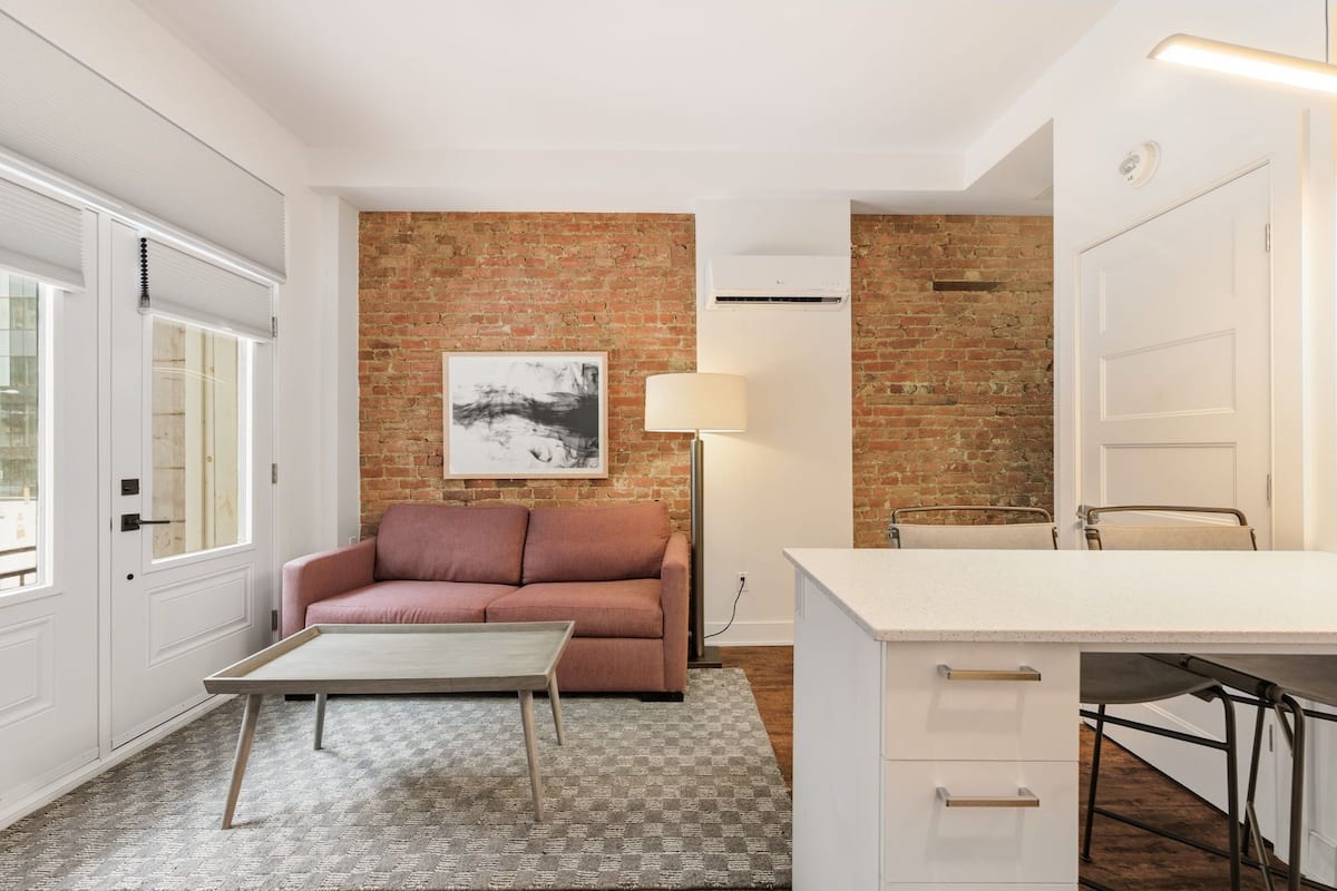 Un espacio habitable moderno con pared de ladrillo, sofá, mesa de centro, lámpara y zona de cocina.