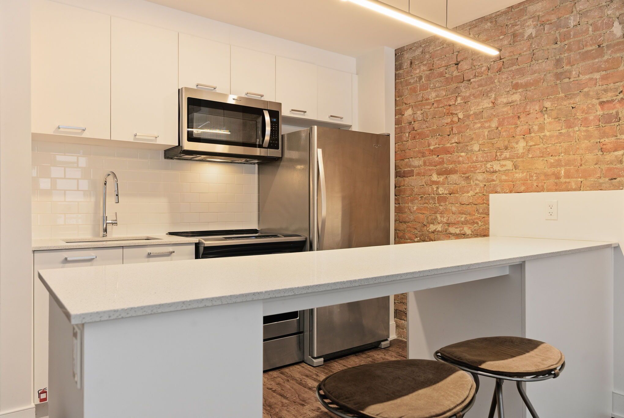 A modern kitchen with white cabinets, stainless steel appliances, a brick accent wall, and a bar area with stools.