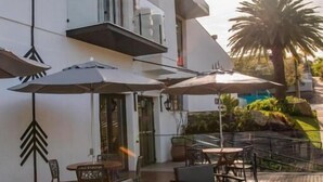 A modern outdoor seating area with a wooden deck, a white building with balconies, and a palm tree in the background. - COROADOS HOTEL E FLATS LTDA (Nova Prata)