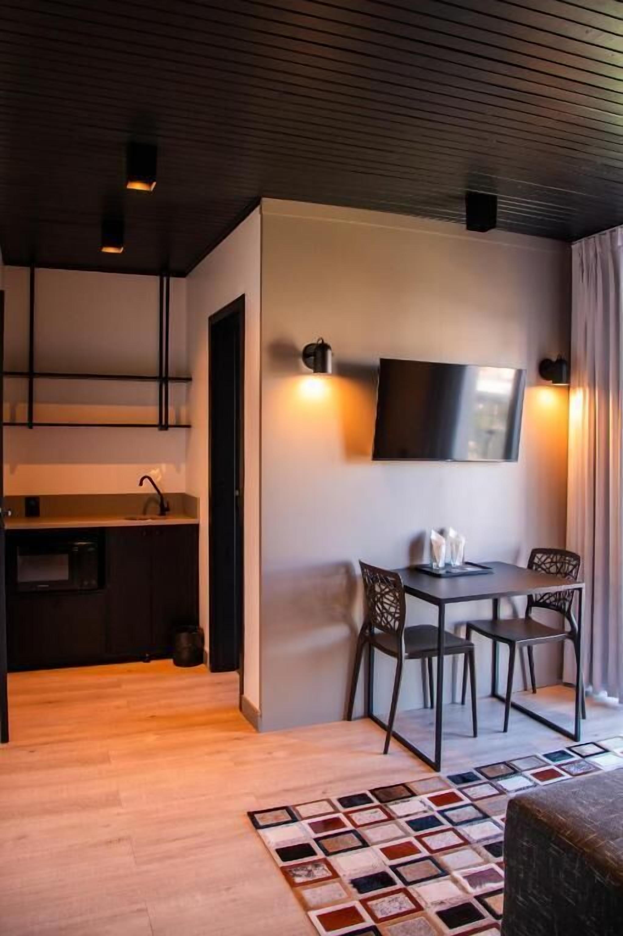 Una cocina moderna con armarios oscuros, una zona de comedor con mesa y sillas, un televisor de pantalla plana en la pared y una alfombra estampada en el suelo.