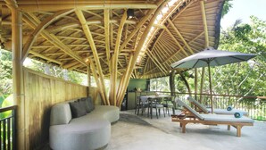 A covered outdoor seating area with a bamboo roof, cushioned seating, and a table with chairs. - Babakan Hill Villas (Karangasem)