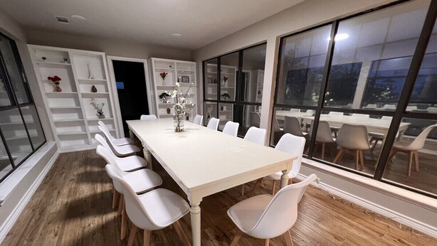 A dining area with a long table and white chairs, wooden flooring, and a view through large windows.