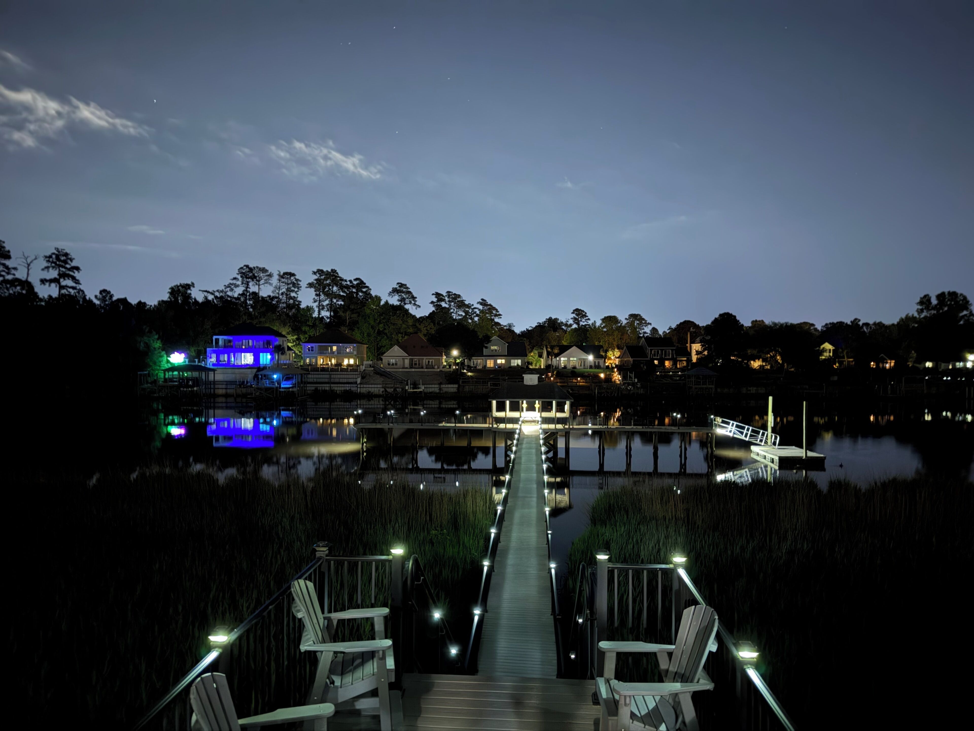 A dock extending into a calm body of water with a row of chairs and a pathway leading to a building with illuminated windows.
