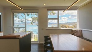 A modern kitchen with a large island, a dining area with a long table, and large windows with ocean views. - Derwent River Holiday Home - Minutes to Hobart (Midway Point)