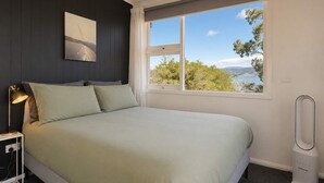 A bedroom with a bed, a nightstand, a lamp, a window with a view, and a standing heater. - Derwent River Holiday Home - Minutes to Hobart (Midway Point)