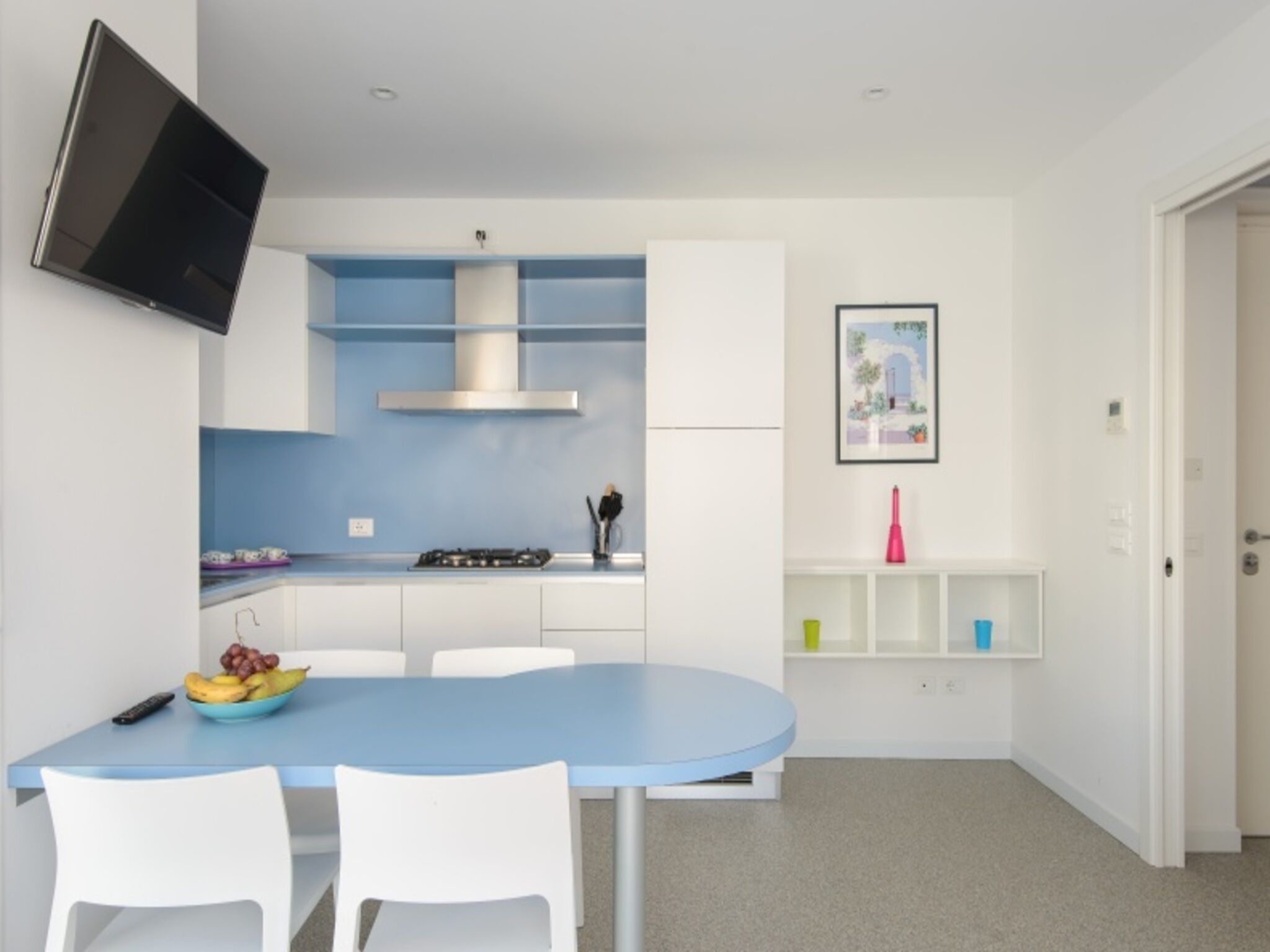 Una cucina moderna con un tavolo blu, sedie bianche, un fornello a gas e una TV a parete.