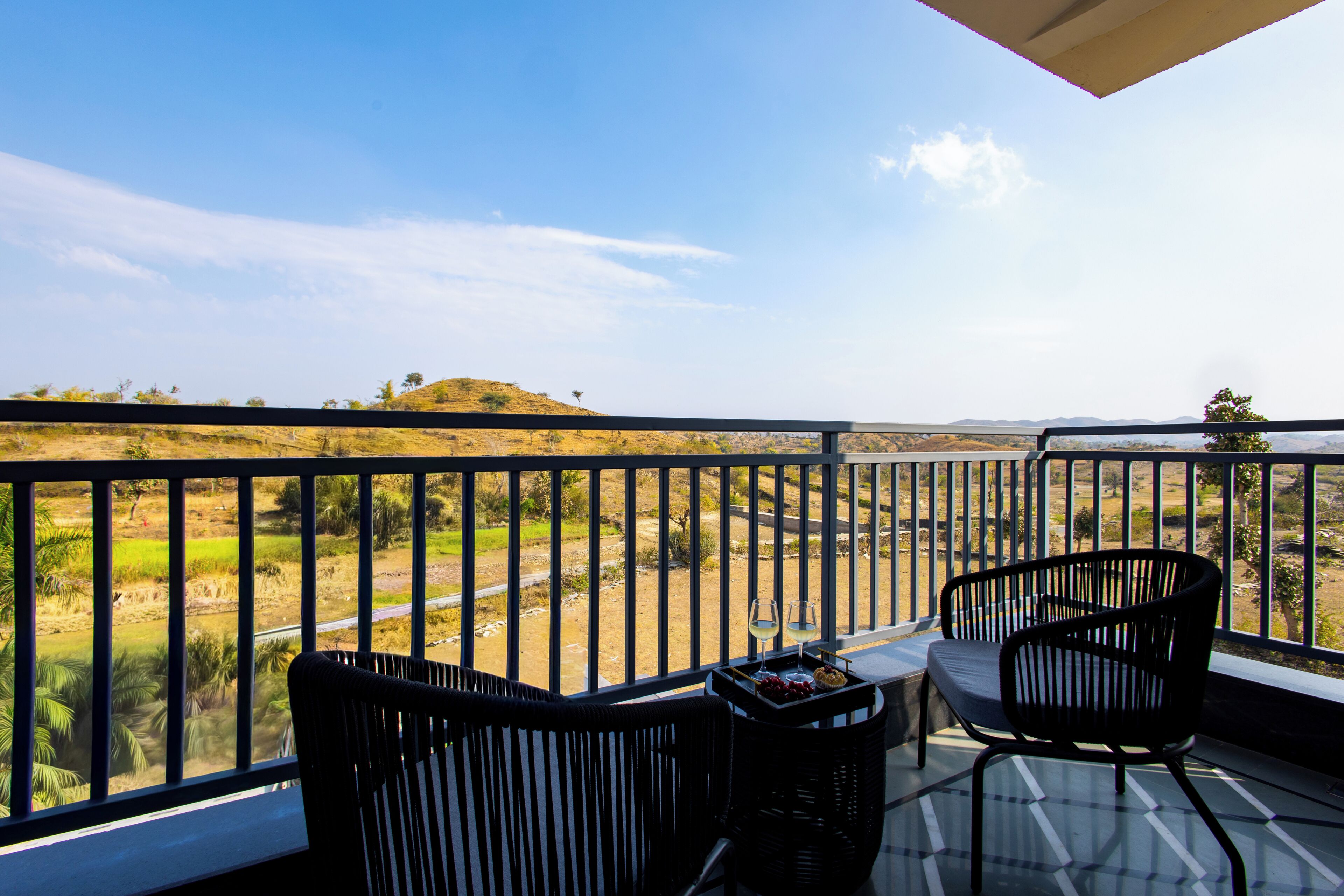 A balcony with two chairs, a table with a tray of snacks and wine glasses, and a view of a landscape with hills and a road.