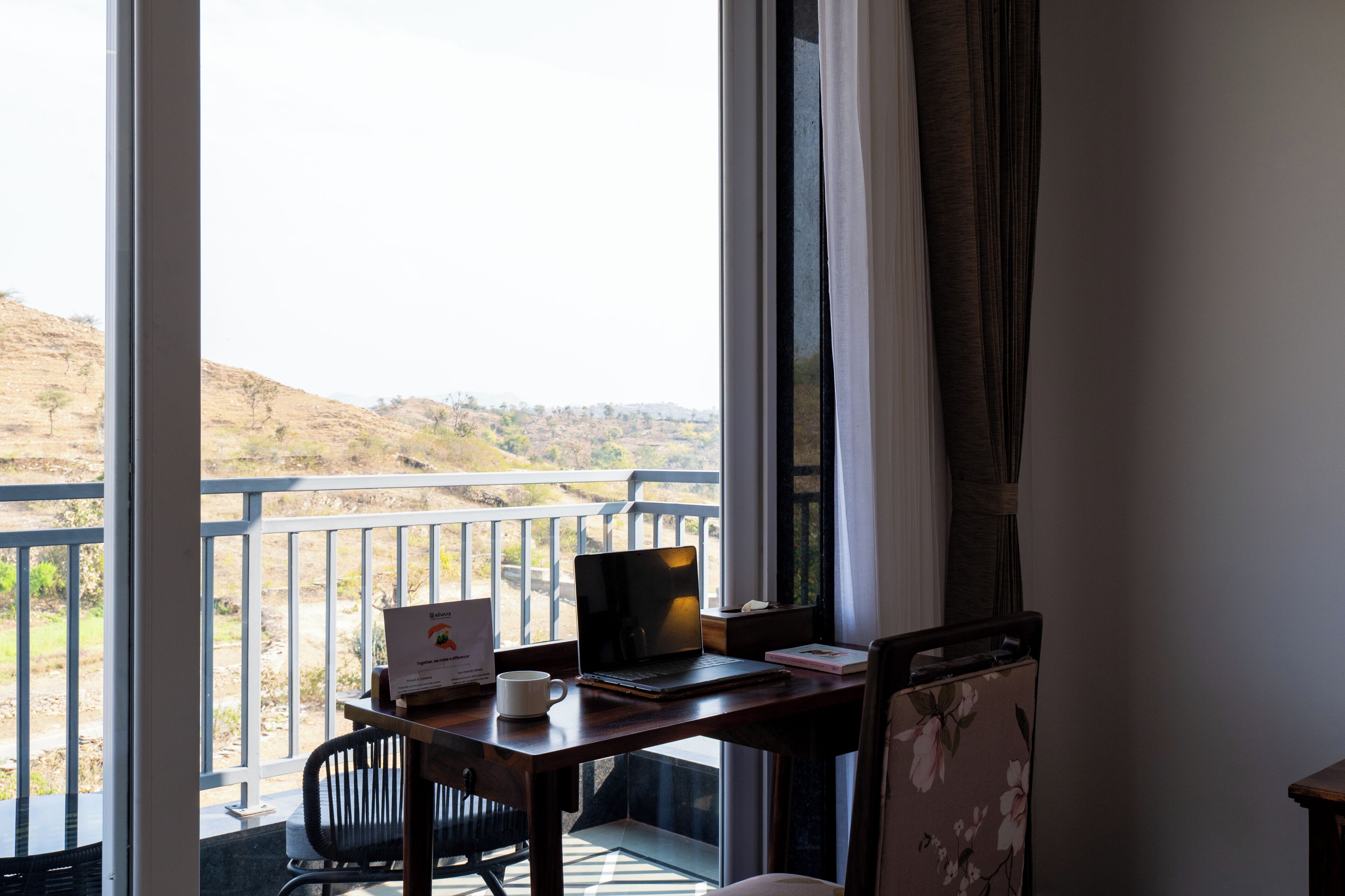 Un balcon avec un ordinateur portable, une tasse et un carnet sur une table en bois, donnant sur un paysage vallonné.