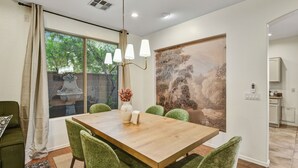 Un comedor con mesa de madera, sillas tapizadas en verde, una araña y un cuadro grande en la pared.