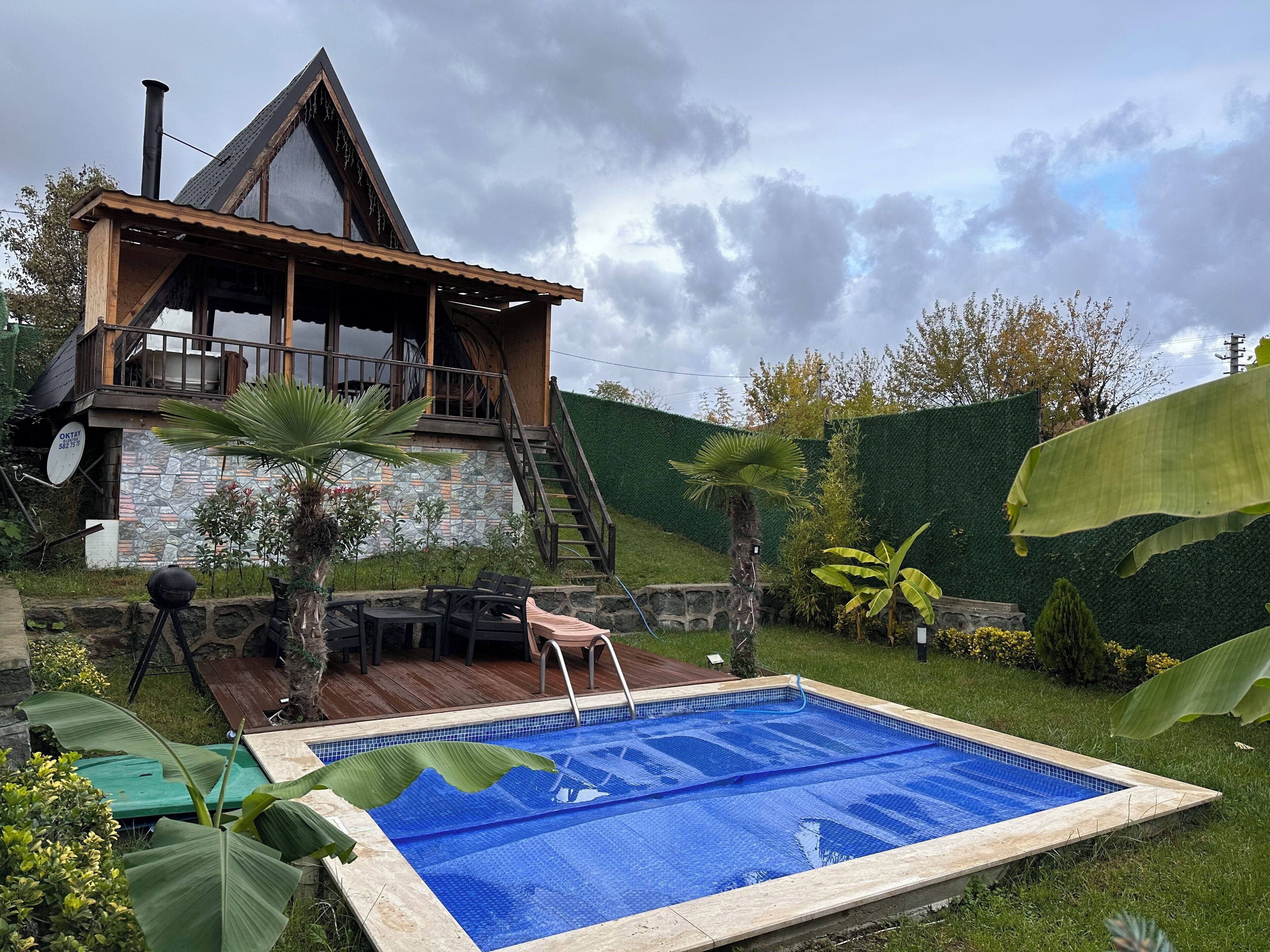 A wooden house with a pool, surrounded by greenery and a stone wall.