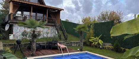 A wooden house with a pool, surrounded by greenery and a stone wall.