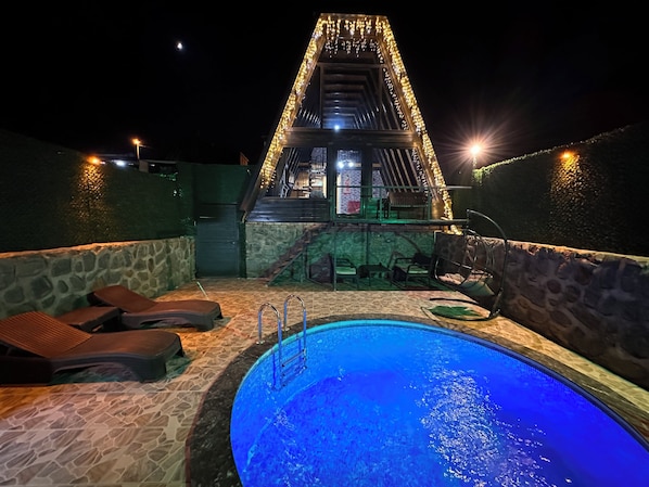A swimming pool with a stone border, lounge chairs, and a staircase leading to a building with string lights.