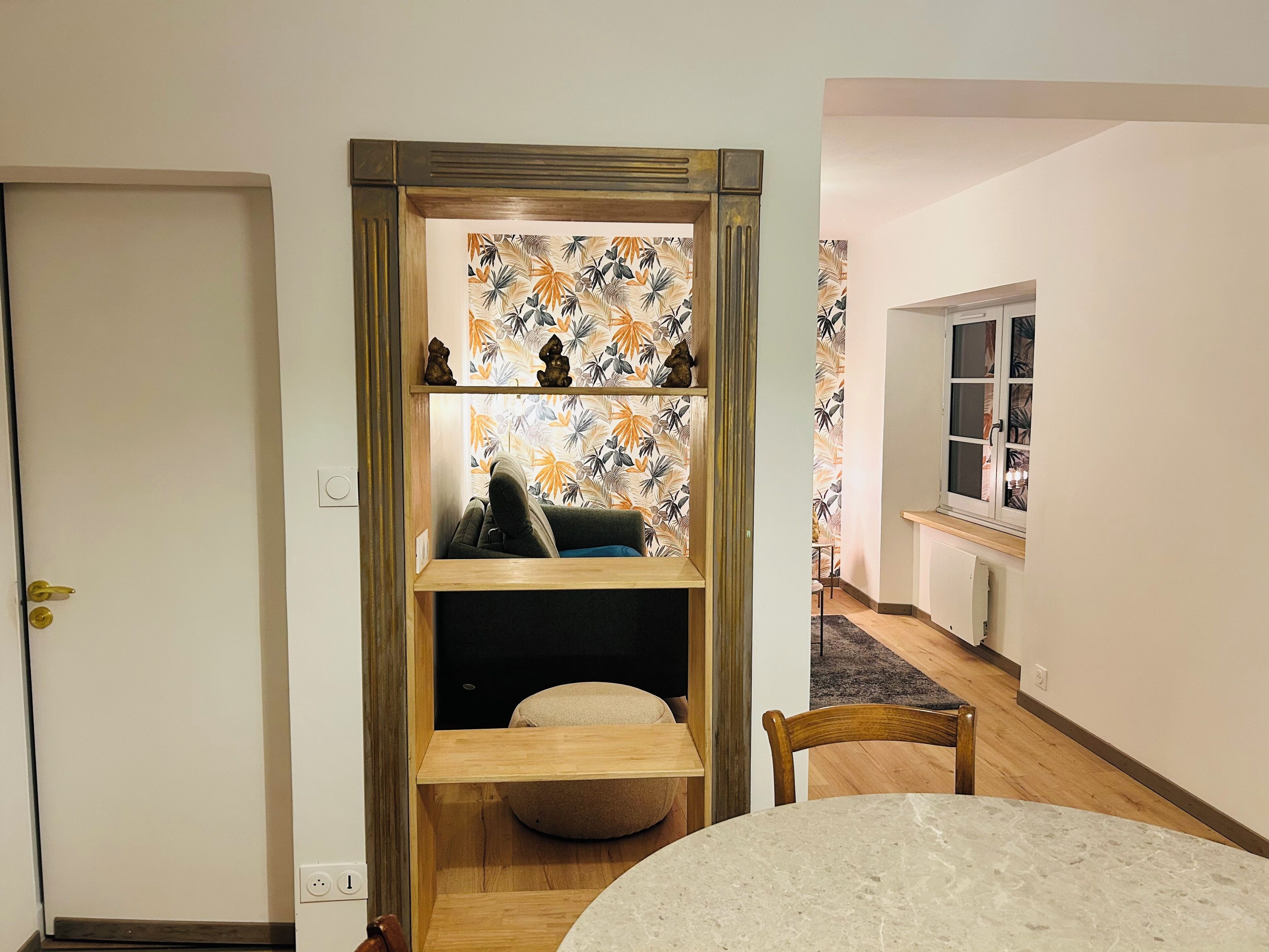 A room with a wooden shelf unit, a round table, and a view into another room with a window.