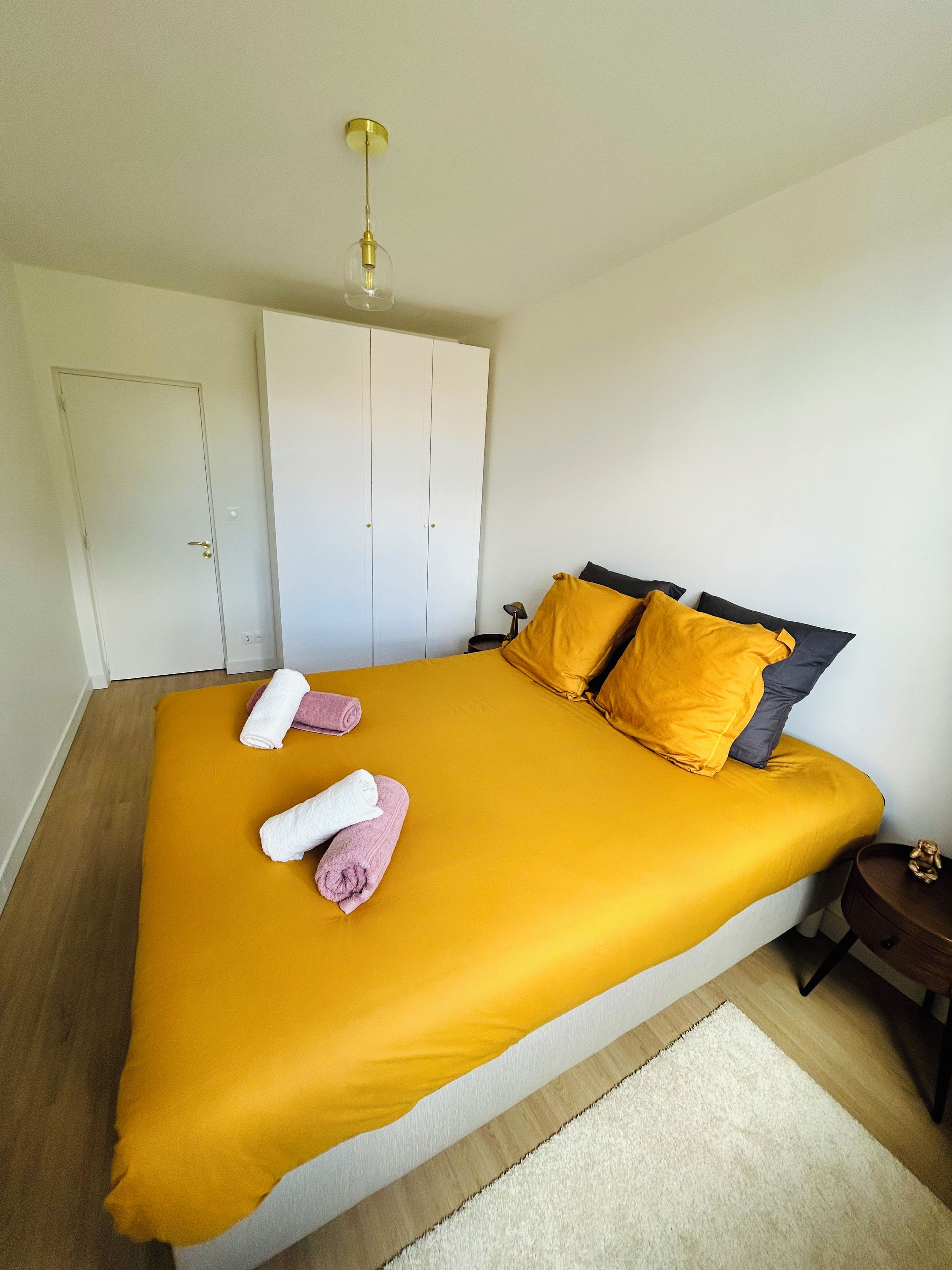 Un dormitorio con cama, almohadas, mesita de noche, armario y una lámpara colgante.