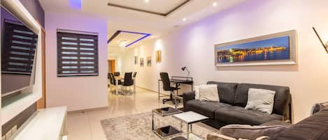 A modern living room with a flat-screen TV, a sofa, a coffee table, and a dining area with chairs and a table.