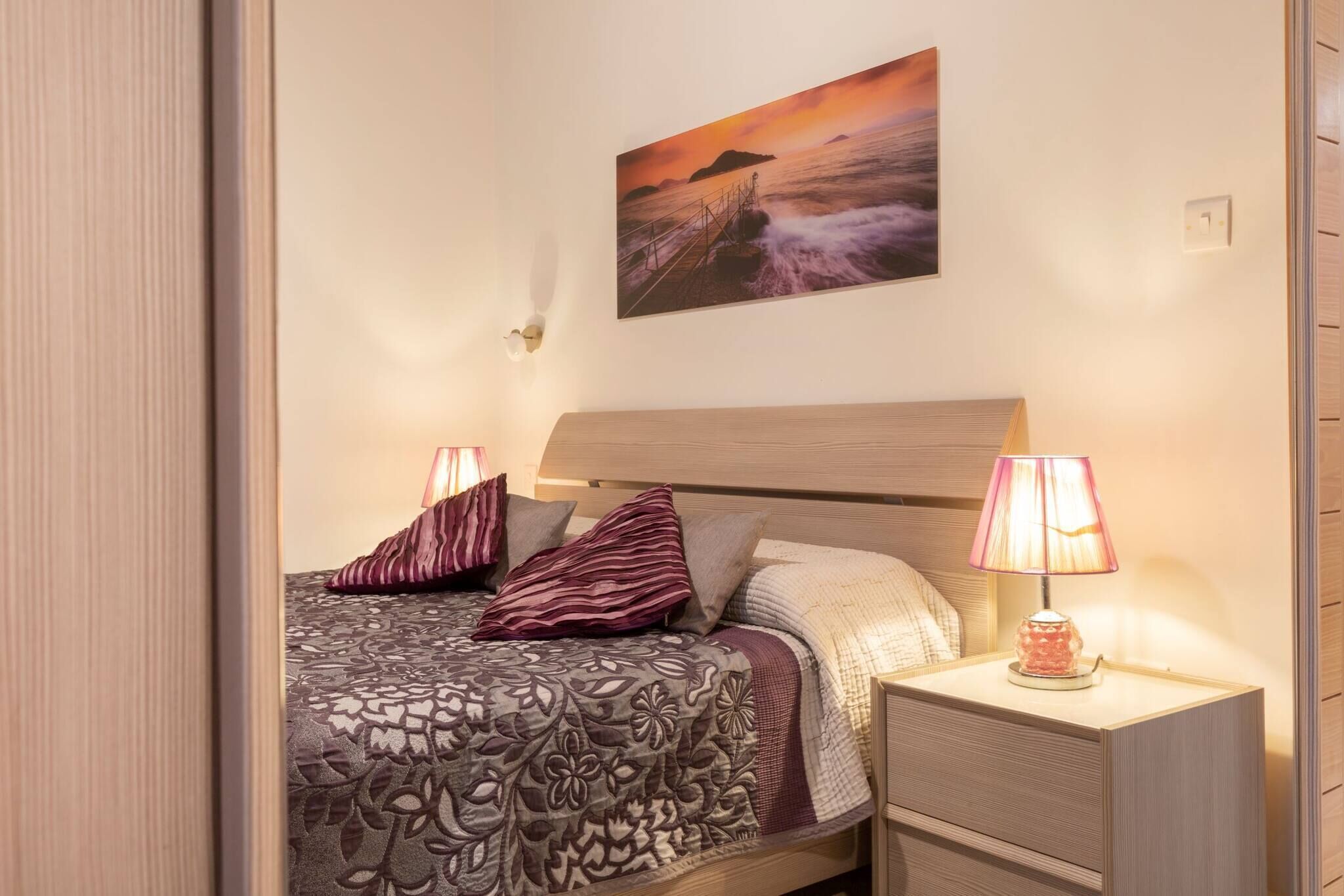 Una cama bien hecha con ropa de cama estampada, una mesita de noche con una lámpara y un cuadro de un atardecer en la pared.