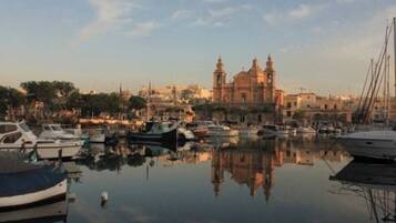 A marina with boats docked, a large church with two domes, and a calm water surface reflecting the structures.