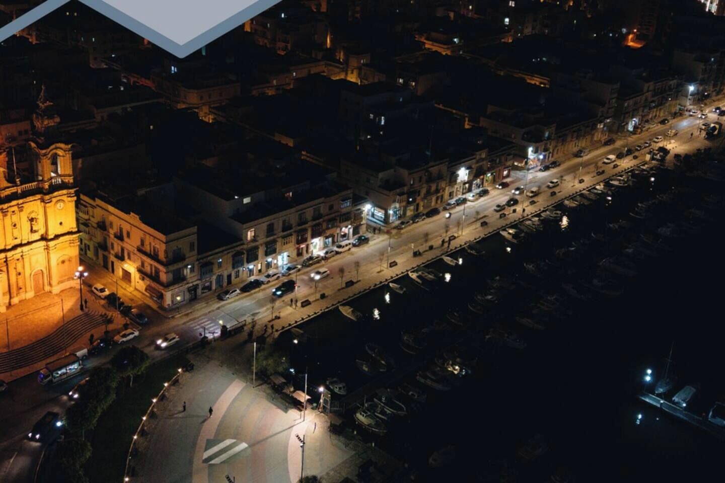 An aerial view of a cityscape at night with illuminated buildings, a prominent church, and a waterfront area with parked boats.